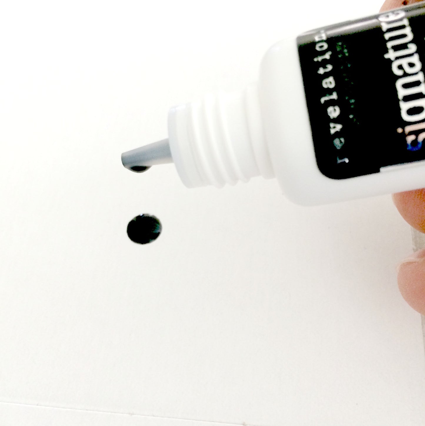 Image of Signature Latex Free Eyelash Extension glue being poured out of bottle. Image shows a drop on white background.